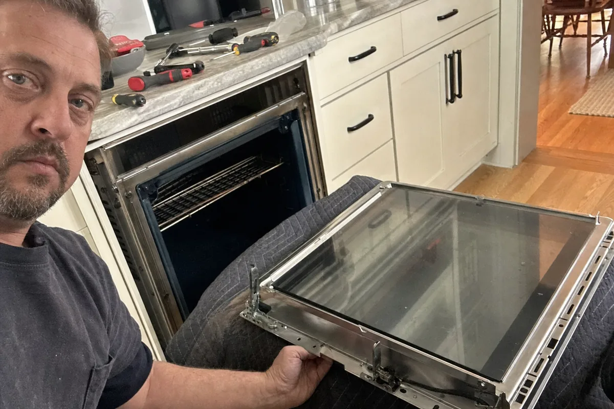 A technician lifting the inner glass panel out of an oven door in a white-cabinet kitchen