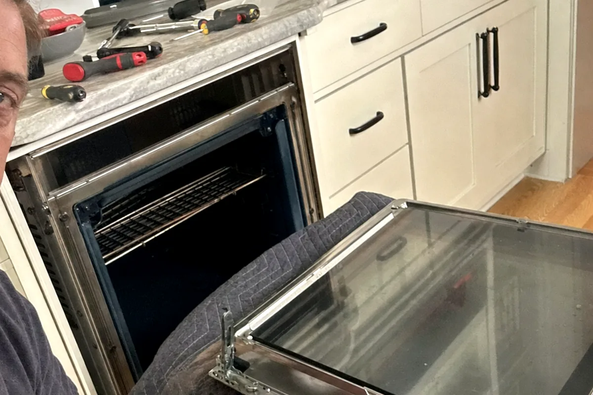An oven door opened on its hinges with the glass panel free of the frame and the hinge arms exposed
